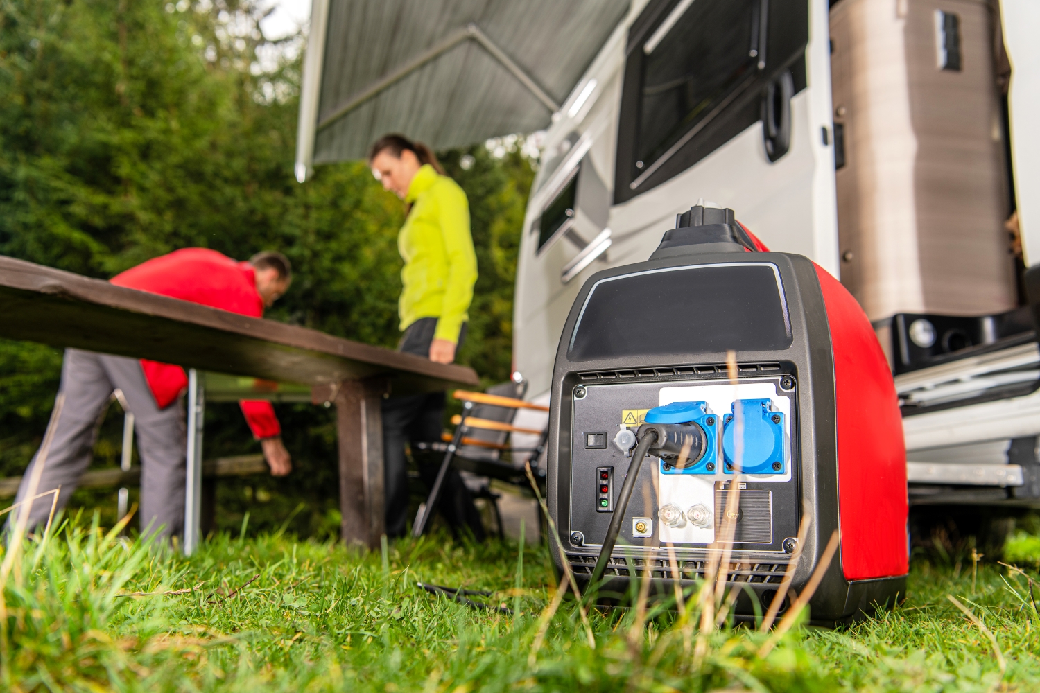 Na pierwszym planie agregat prądotwórczy jako generator prądu dla kampera. W tle dwoje ludzi i kamper